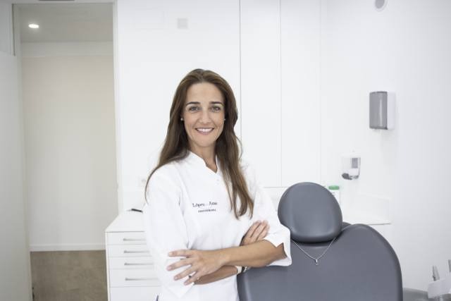 Odontóloga con uniforme blanco y brazos cruzados junto al sillón dental en gabinete clínico